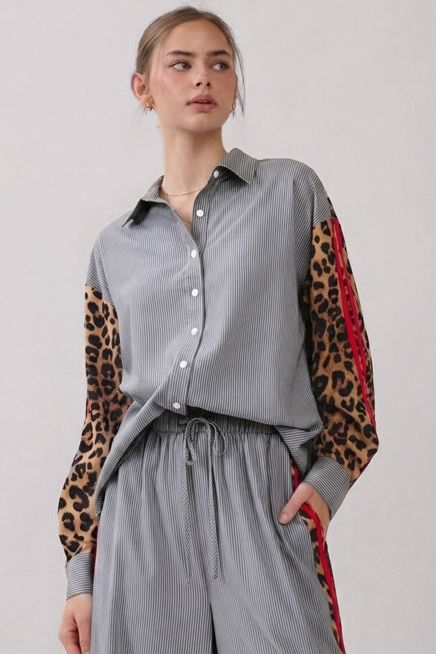 A woman wearing a gray pinstriped button-up shirt with leopard-print sleeves and red side panels, paired with matching gray pinstriped trousers, against a light gray studio background.