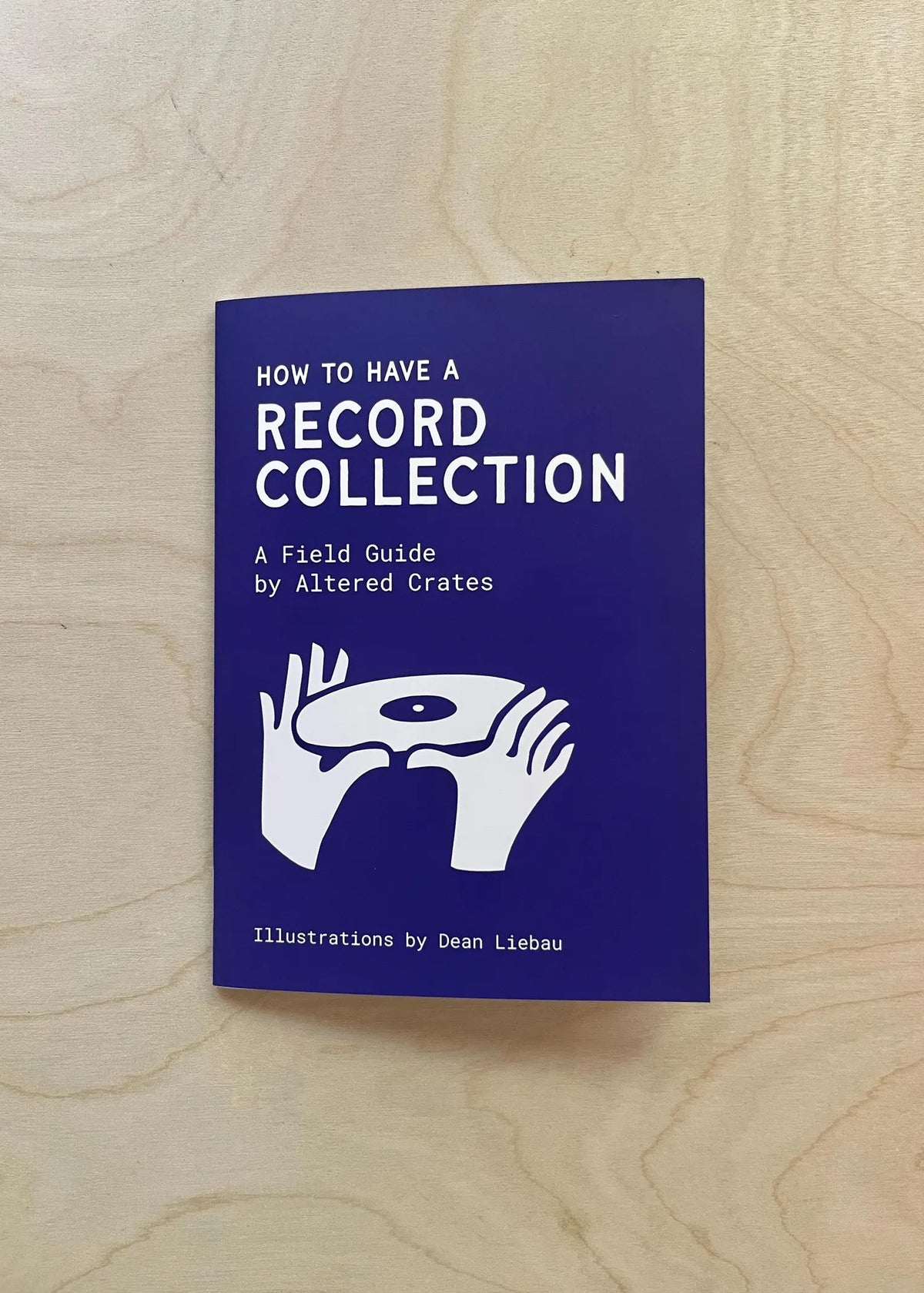 A book titled 'HOW TO HAVE A RECORD COLLECTION' by Altered Crates, featuring a dark blue cover with white text and a graphic of hands holding a vinyl record, placed on a light wood grain surface.